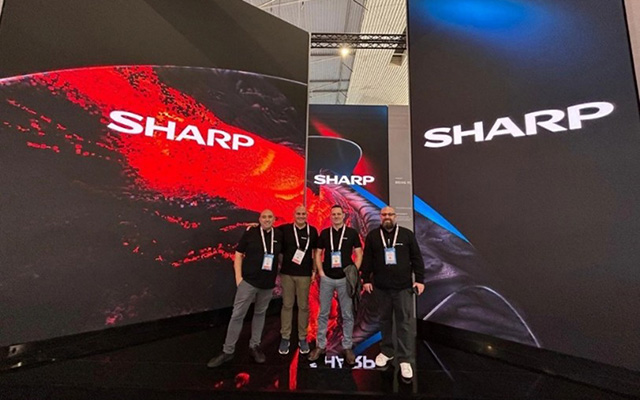 four men in black polo shirts stand in front of three large LED screens showing the Sharp logo